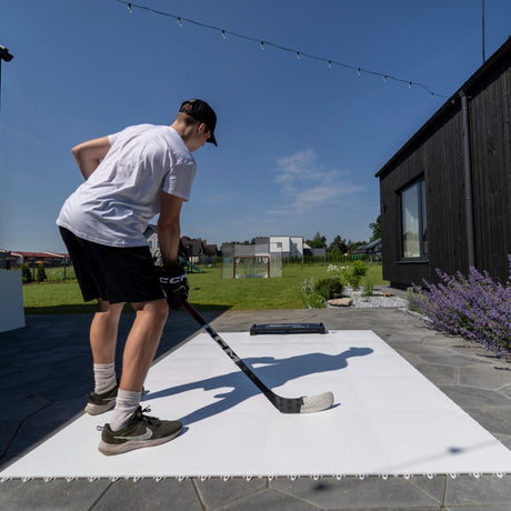 En spelare tränar skott och passningar på ett hockeygolv utomhus, med skottramp och hockeymål i bakgrunden.