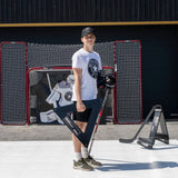 En man står med en extrem hockeypassare i hand, klädd i t-shirt och keps, med hockeymål i bakgrunden.