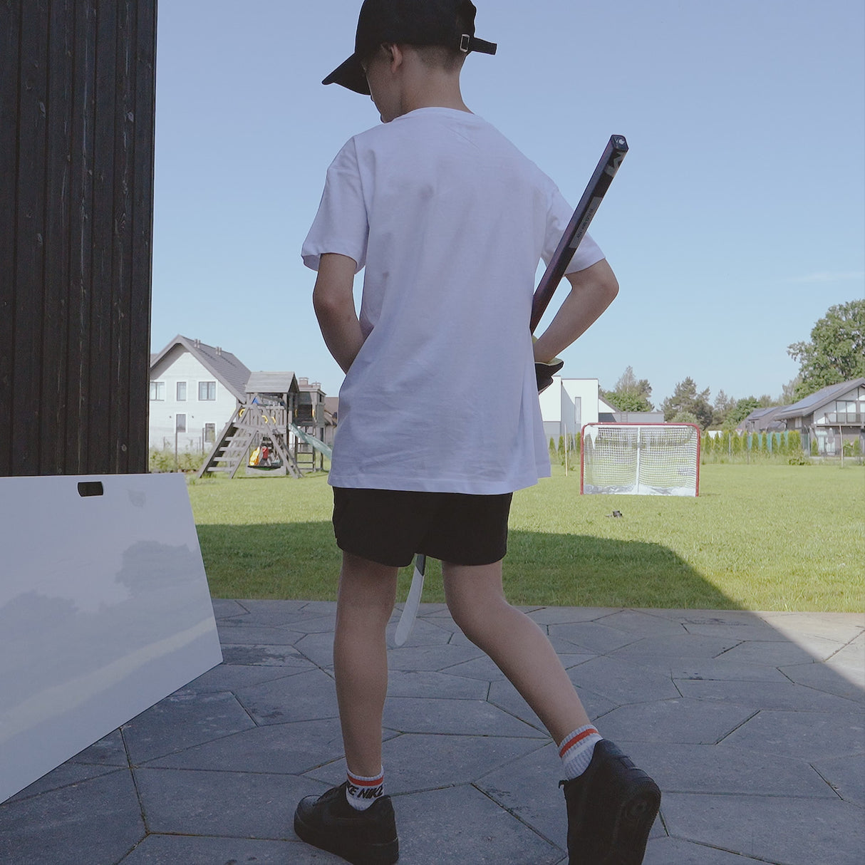 Ung pojke i vit t-shirt och shorts som bär en hockeyklubba, går mot en skottplatta i en trädgård.